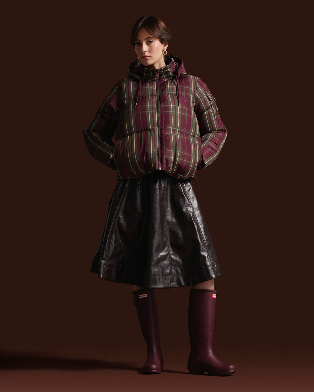 A model stands against a dark brown studio background wearing a hooded puffer jacket with a burgundy, green, and tan plaid pattern, a black faux-leather skirt, and dark burgundy rain boots.