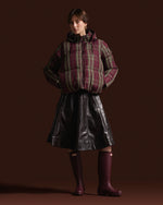 A model stands against a dark brown studio background wearing a hooded puffer jacket with a burgundy, green, and tan plaid pattern, a black faux-leather skirt, and dark burgundy rain boots.
