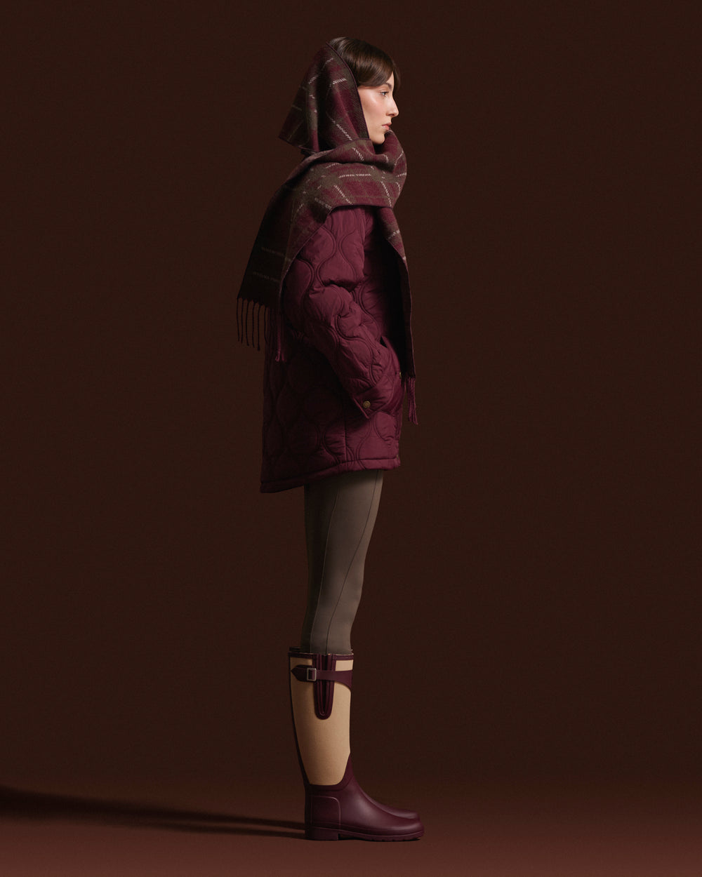 A model in profile wearing a dark plum quilted puffer coat, a matching plaid scarf, olive green leggings, and dark plum rain boots against a solid dark brown studio background.