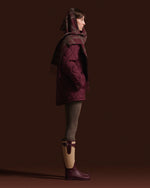 A model in profile wearing a dark plum quilted puffer coat, a matching plaid scarf, olive green leggings, and dark plum rain boots against a solid dark brown studio background.