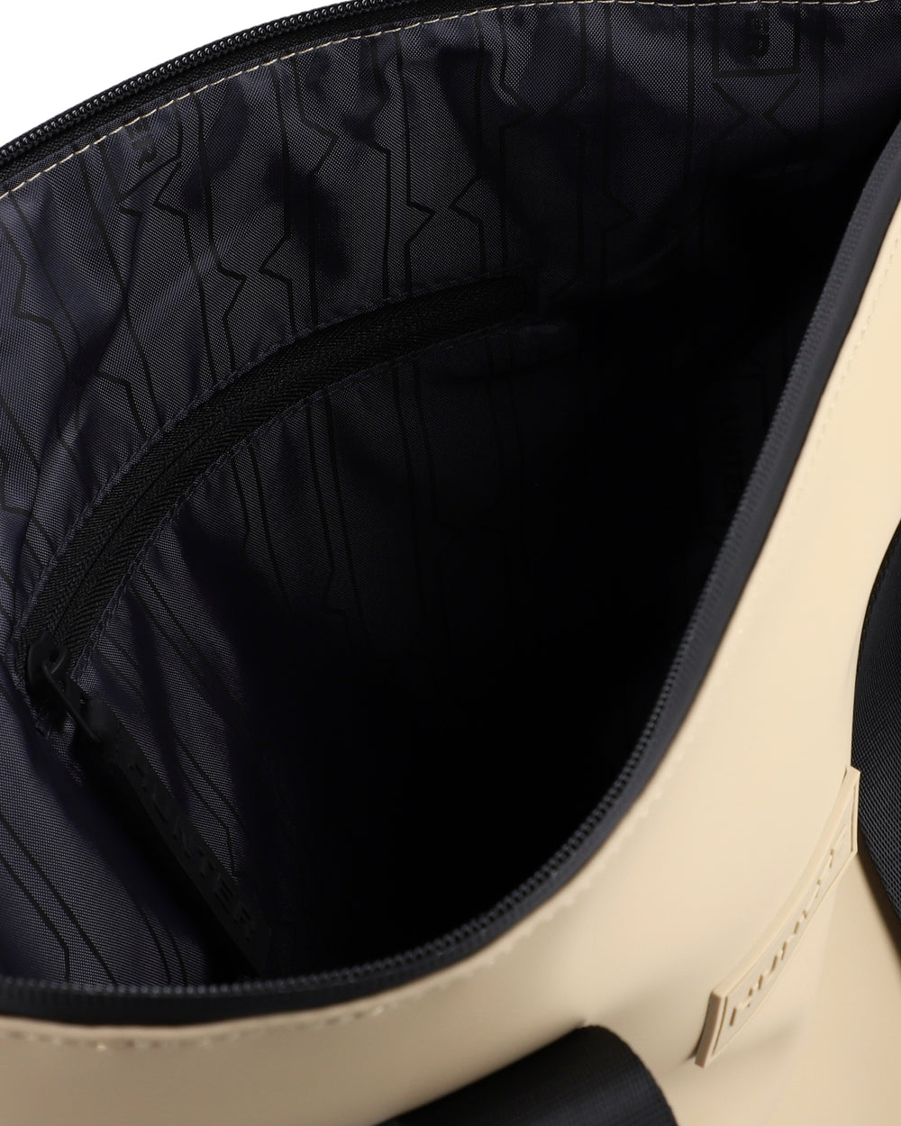 Close-up of the interior of a beige tote bag, showing a black zippered pocket lined with black patterned nylon and a small beige brand tag on the side.