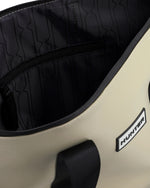 Close-up of a Hunter duffel bag showing the black interior lining with a repeating logo pattern, a black zipper, and a black webbing handle on a light beige exterior with a white and black Hunter brand patch.