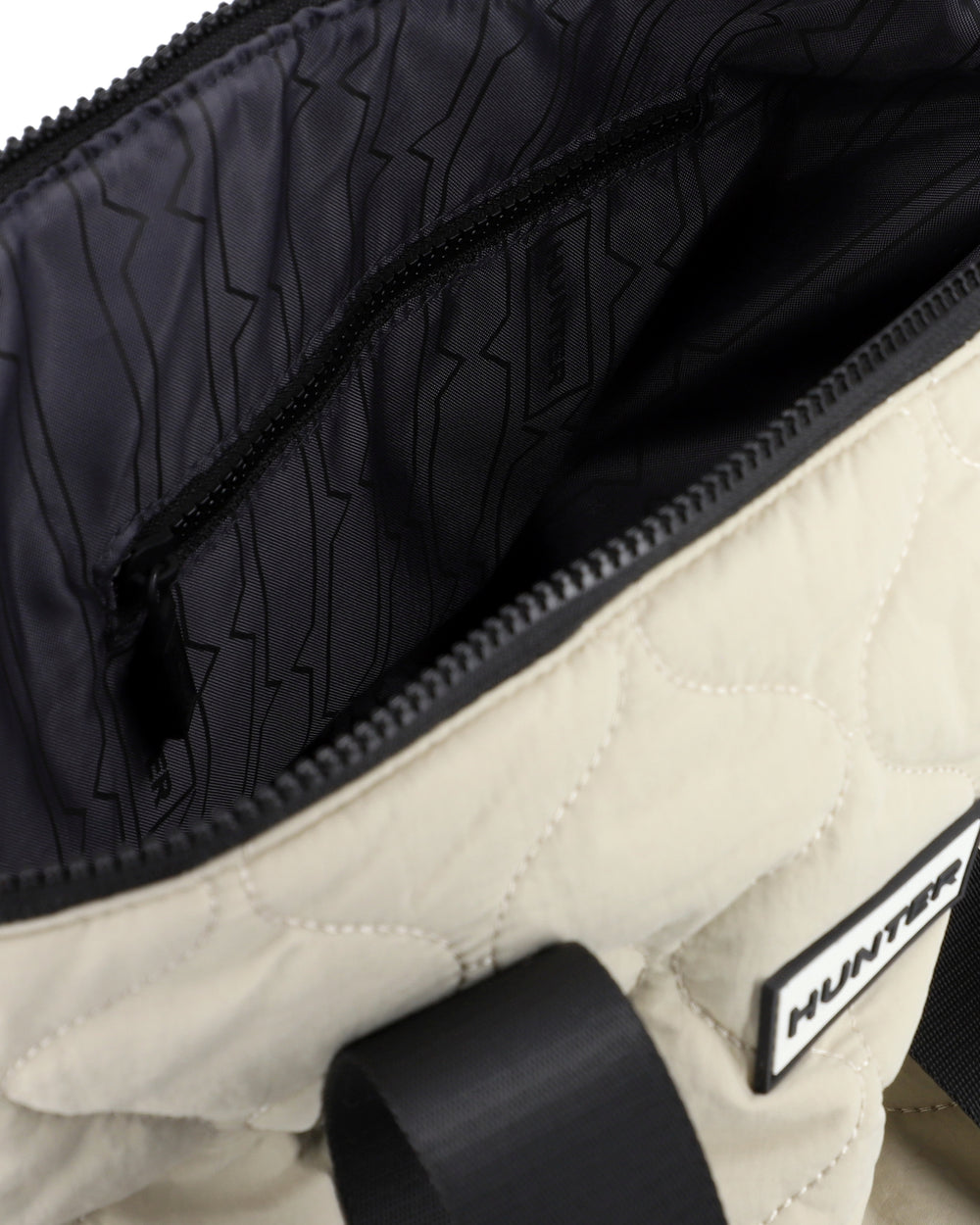 Close-up of a quilted off-white and black bag, showing the interior lining with a zigzag pattern, a black zipper pocket, and a black brand label reading HUNTER.