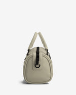 A light beige faux-leather handbag with a small dome shape, featuring dual top handles, a vertical black strap detail on the front, and silver-tone hardware, set against a plain white background.